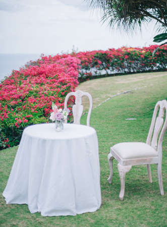 A small round table with two white chairs in a garden with beautiful red roses on a sunny dayの写真素材