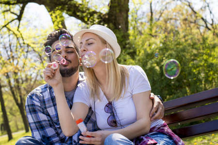 A young attractive couple having fun while blowing bubbles together on a park benchの写真素材