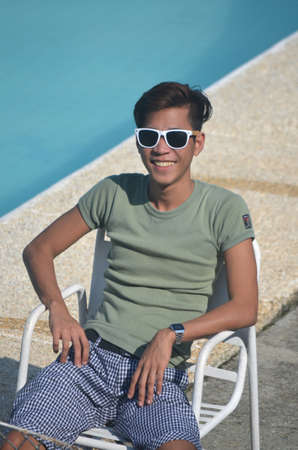 A vertical shot of a male wearing a grayish green shirt and sunglasses sitting on a chair by the poolの写真素材