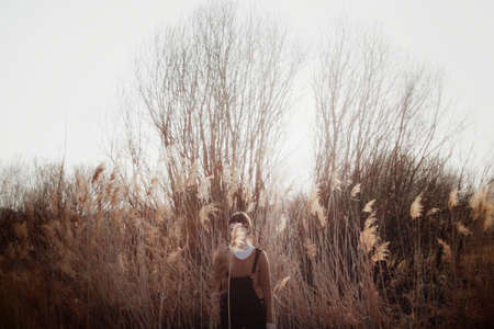 A sad female whose face is covered with grass standing in the tall dry bushes in the evening