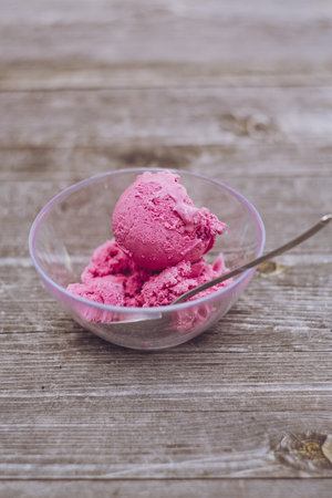 A clear glass bowl of delicious strawberry ice cream and a spoon on a wooden surface