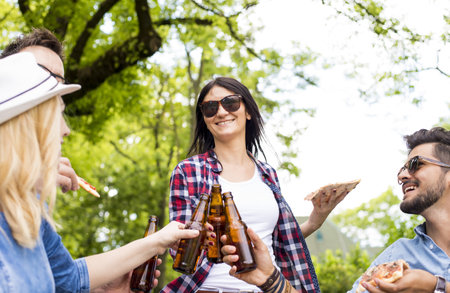 A group of young friends drinking beers and eating pizza on a park bench togetherの写真素材