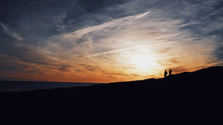 A wide shot of silhouettes of two people walking on a hill with the sun shining in the background