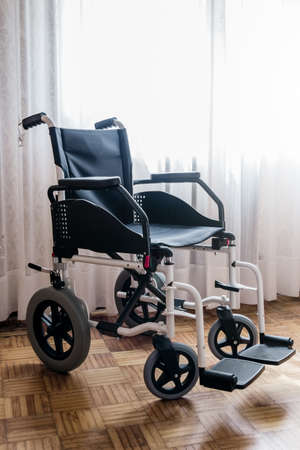 A vertical shot of an empty wheelchair in a room with wooden floors and bright white drapesの写真素材