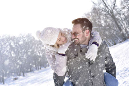 A happy caucasian couple enjoying a piggyback ride in a snowy field, having fun and enjoying each other's company during daylightの写真素材