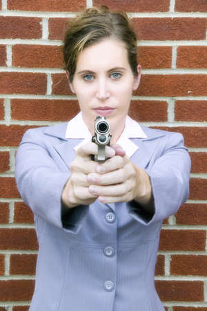 A vertical shot of a young Caucasian woman holding a gun with a brick wall on the background