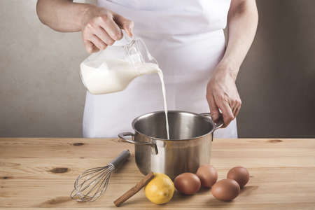 A high angle shot of a chef pouring milk into a pot set on a wooden table next to some eggs and a lemonの写真素材
