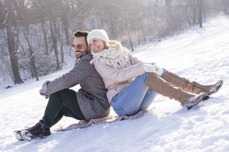 A happy caucasian couple in a snowy field sitting on the snow and enjoying each other's company during daylightの写真素材