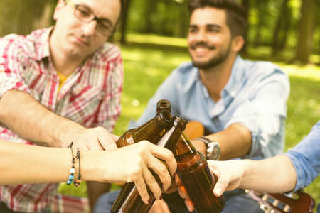 A closeup of a group of caucasian friends toasting with their beer bottles, in a park during daylightの写真素材