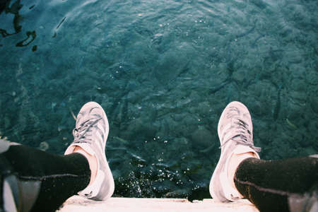 An overhead closeup shot of a person wearing sneakers sitting above the body of waterの写真素材