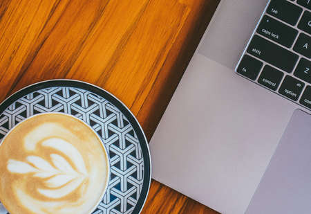 A closeup shot of a beautifully made cup of cappuccino next to a laptop on a wooden surfaceの写真素材