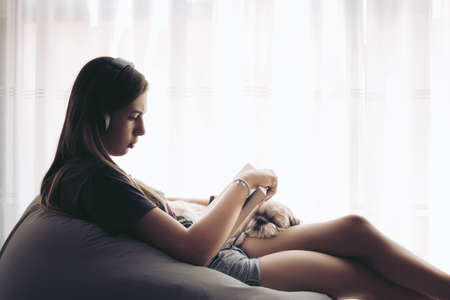 A young female reading a book using headphones with a dog on her lapの写真素材