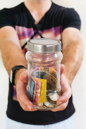 A selective closeup shot of a male holding a glass jar filled with Australian moneyの写真素材