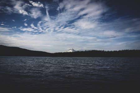 A beautiful silhouette shot of trees by the body of water near a mountain under a cloudy blue skyの写真素材