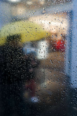 A vertical shot of a glass with raindrops and a person with a yellow umbrella in the backgroundの写真素材