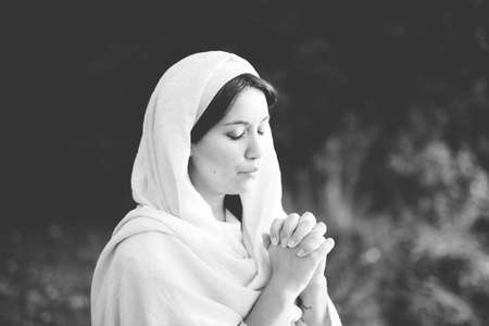 A grayscale shot of a female wearing a biblical gown and praying while her eyes are closed