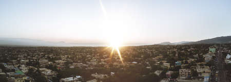 A panoramic shot of city buildings with trees and the sun shining in a blue sky in the backgroundの写真素材