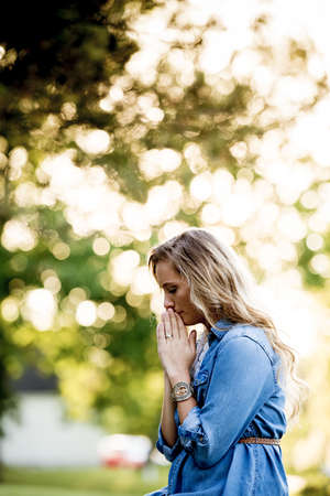 A woman wearing a blue dress and praying in a garden under sunlight with a blurry background