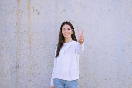 An attractive brunette with a happy smile wearing a white sweater showing the peace sign isolated on a white backgroundの写真素材