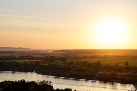 A high angle shot of a river in the middle of the grassy field and trees with a bright sky in the backgroundの写真素材