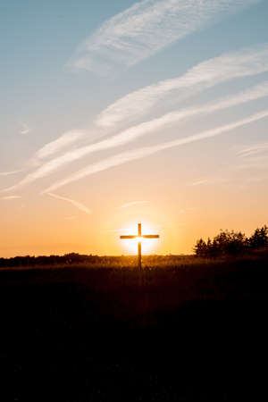 A vertical shot of a cross in a grassy field with the sun shining in the backgroundの写真素材