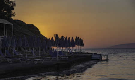 A beach resort with a lot of parasols and folding chairs near the sea under the sunset skyの写真素材