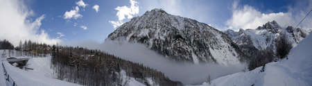 A panoramic shot of a rocky mountain covered with snow and fog during winter with a blue sky in the...
