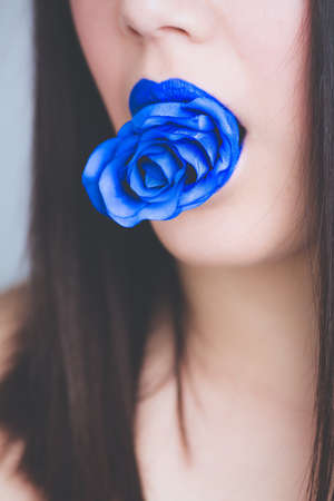 A closeup shot of a dark-haired female with a blue lipstick biting a blue rose - great for a fashion blog articleの写真素材