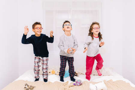 The three cheerful children with glasses jumping and playing on the bedの写真素材