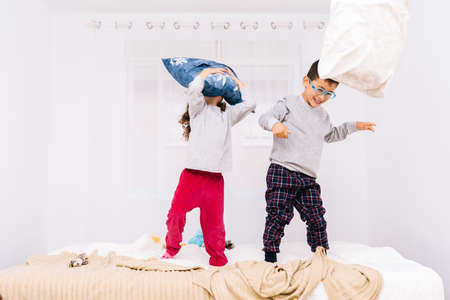 The cheerful children with glasses jumping and playing with pillows on the bedの写真素材