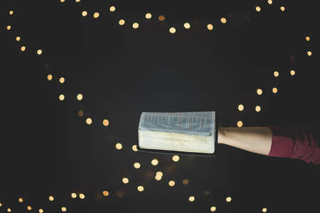 A closeup shot of a person holding an open bible with blurred lights in the backgroundの写真素材