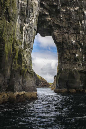 A vertical shot of a beautiful natural arch in the sea with a blue cloudy sky in the backgroundの写真素材