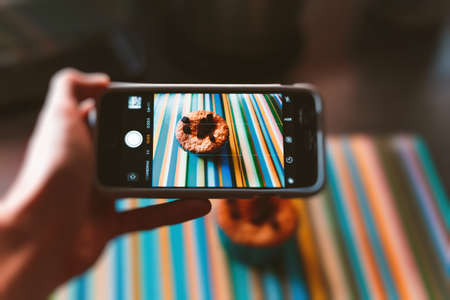 A horizontal closeup shot of a person with a smartphone taking a picture of a muffin with chocolate chip toppingの写真素材