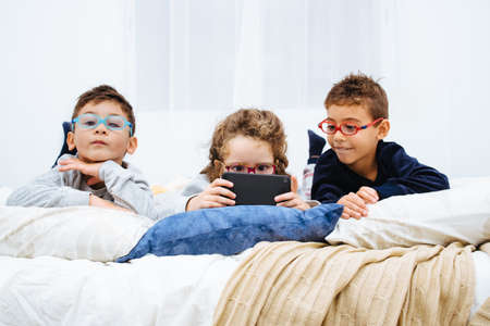 The cheerful three children with glasses playing on the phone on the bed