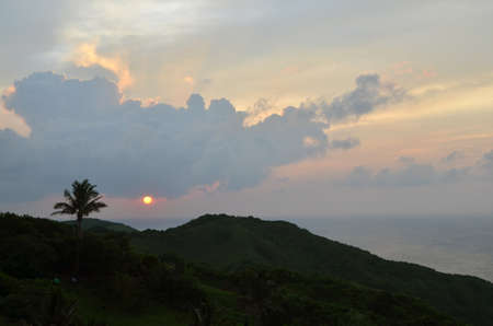A high mountain covered with greenery and palm trees under a cloudy sky during sunsetの写真素材