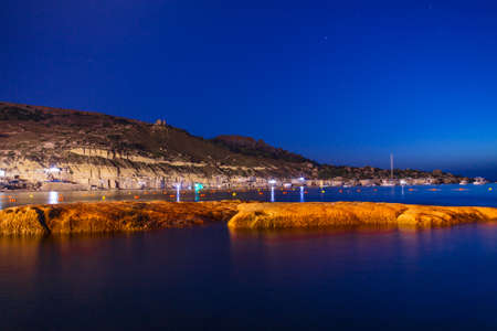 A beautiful shot of rocky hills with lights near the sea under the beautiful blue skyの写真素材
