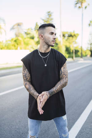 A vertical shot of a cool young man with tattoos wearing a black t-shirt standing on the streetの写真素材