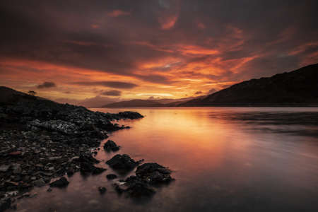 A wide shot of black rocks by the water surrounded by mountains during dawn