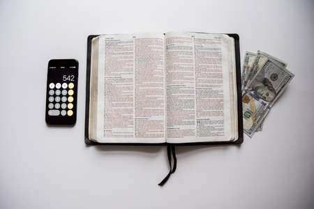 An overhead shot of an open bible in the middle of money and a smartphone on a white surfaceの写真素材