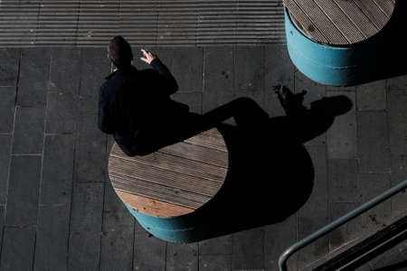 A high angle shot of a lonely male sitting on a round bench and his shadow smoking a cigarette on the streetの写真素材