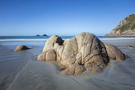 A beautiful shot of a rock formation on a sandy shore by the water surrounded by a cliff under a...