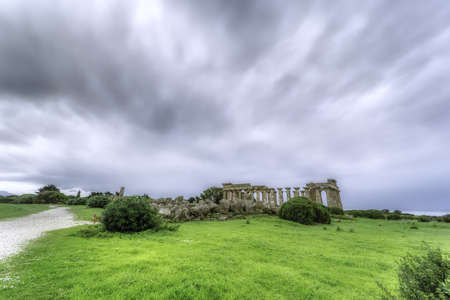 A wide shot of isolated abandoned building ruins in a green field under the dark cloudy skyの写真素材