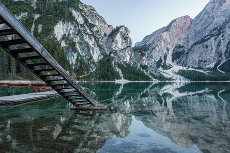 A beautiful scenery of high rocky mountains reflected in Braies lake with wooden stairs near the pier in Italyの写真素材