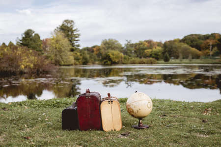 A closeup shot of old suitcases on a grassy field near a desk globe with blurred water in the backgroundの写真素材