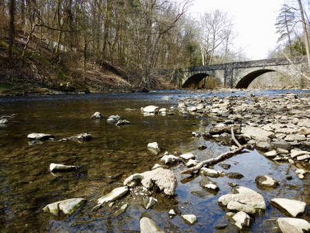 A wide shot of a river under the bridge surrounded by bare trees and a lot of rocks