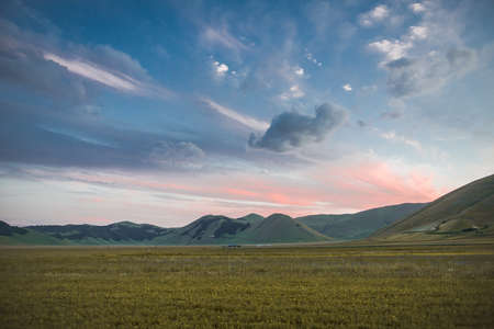A wide shot of beautiful green mountains in a grass field under the colorful cloudy sky in Italyの写真素材