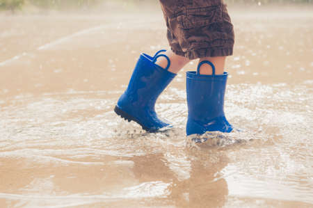 A closeup shot of the legs of a boy in blue rubber boots splashing in a puddle