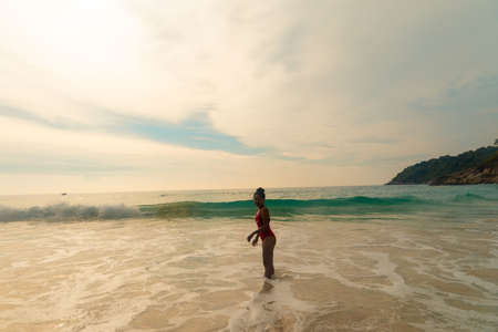 A beautiful shot of a female wearing a red swimsuit standing at the ocean shore during sunsetの写真素材
