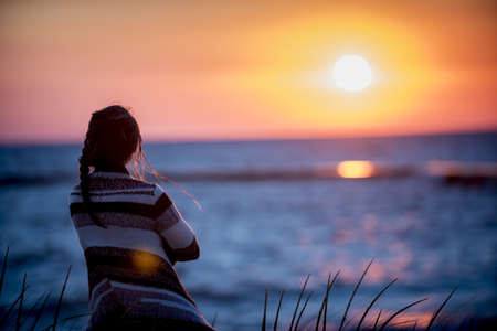 A shallow focus shot from behind of a female looking at the sea at sunsetの写真素材