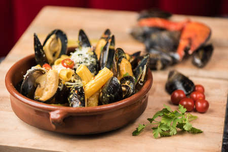 A closeup of mussel and macaroni with cheese in a bowl near vegetable leaf and tomatoes on a wooden...